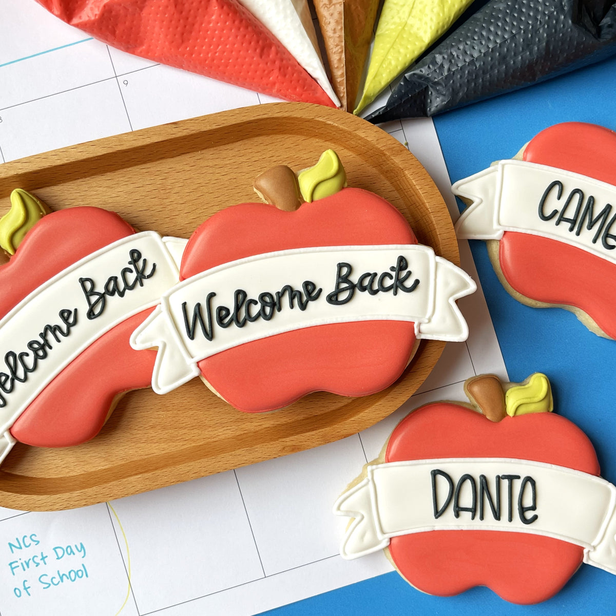 Red apple-shaped cookies with white icing banners reading welcome back and dante decorated with yellow-green leaves using apple banner plaque cookie cutter