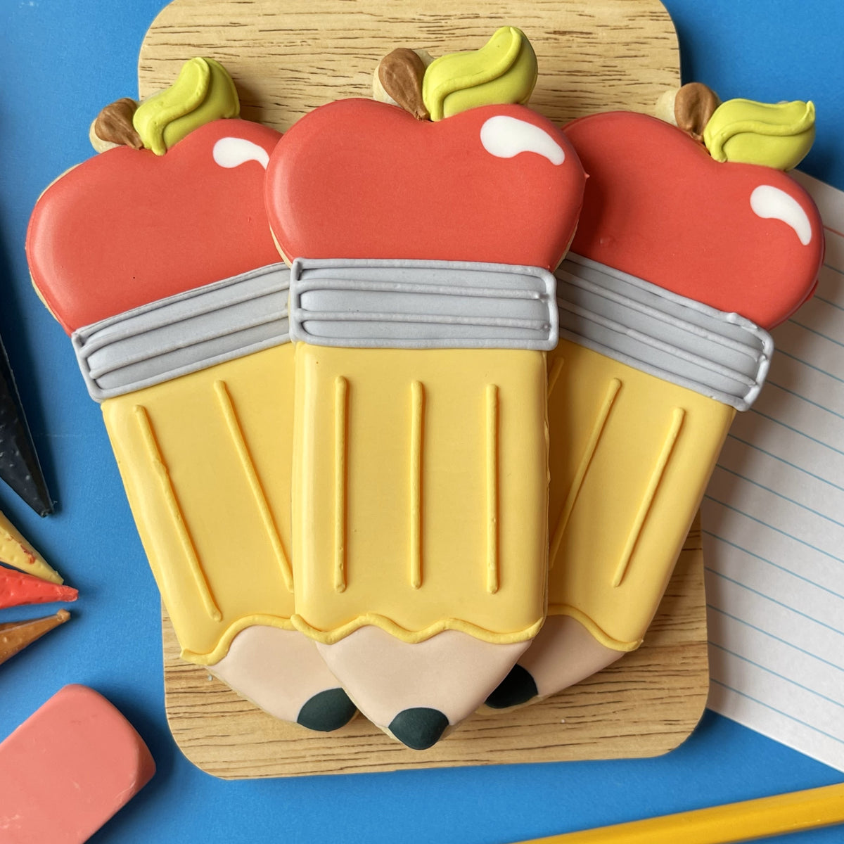 Three red apple shaped cookies with yellow pencil bodies and green leafy stems made using an apple pencil cookie cutter