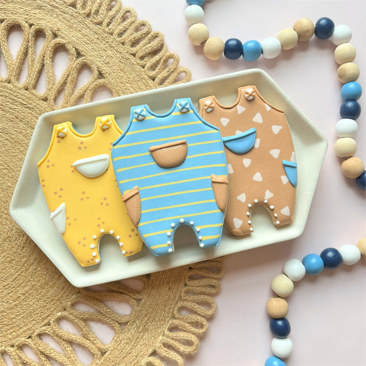 Three baby overall cookie cutter cookies decorated with yellow brown dots blue stripes and beige white hearts on plate