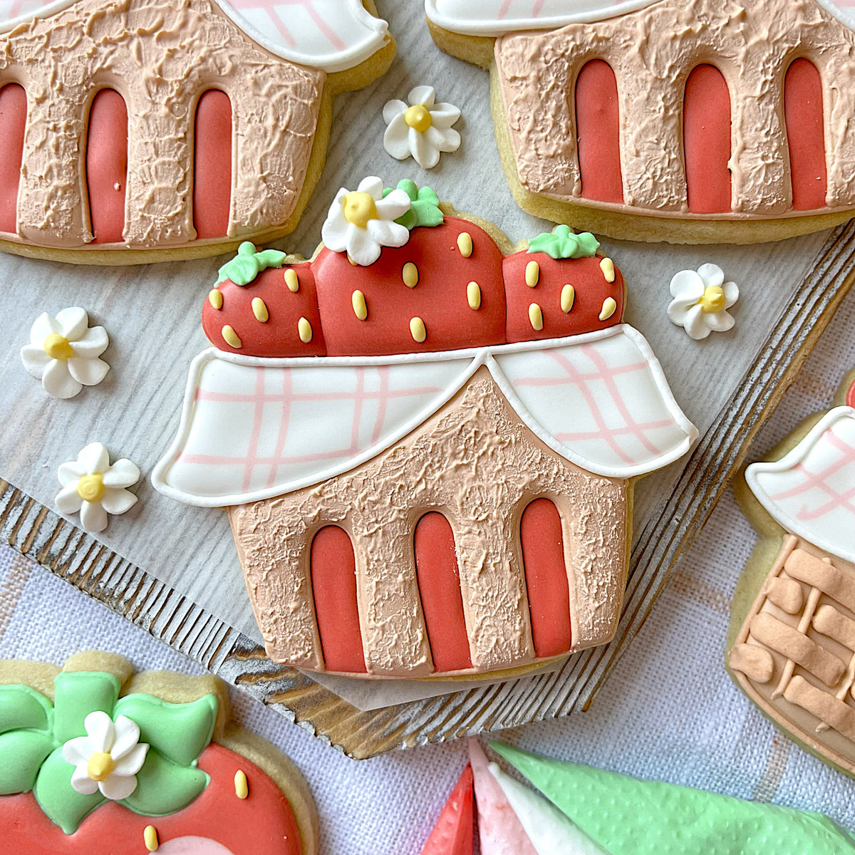 A red strawberry-themed cookie shaped like a house with pink and white checkered icing roof for berry basket cookie cutter
