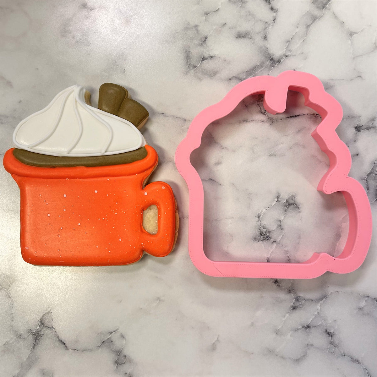 Orange cookie shaped like a mug with white and brown icing next to a pink silicone mold from campfire mug cookie cutter