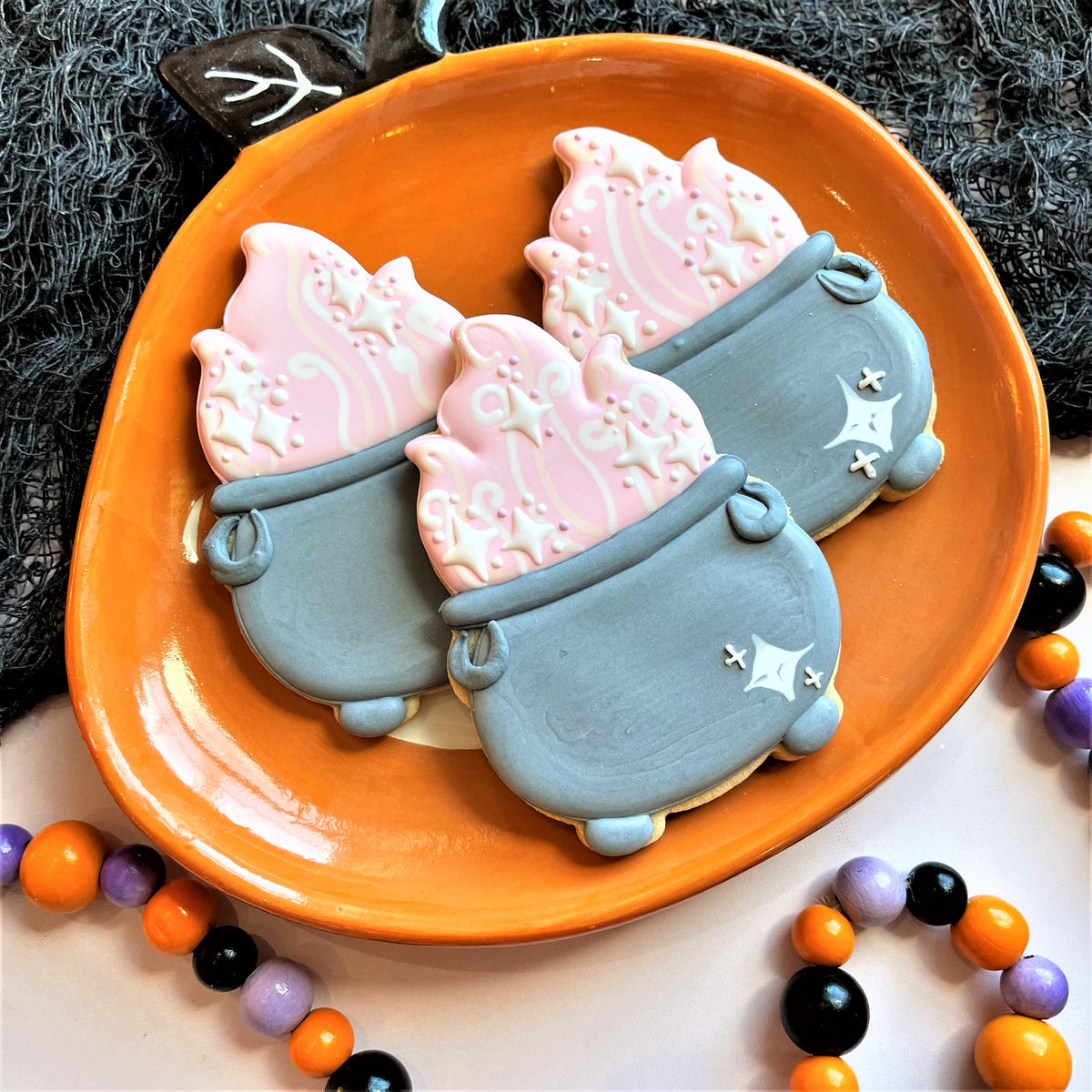 Three gray witch cauldron cookies with pink star icing on an orange pumpkin plate for cute witch decorating class
