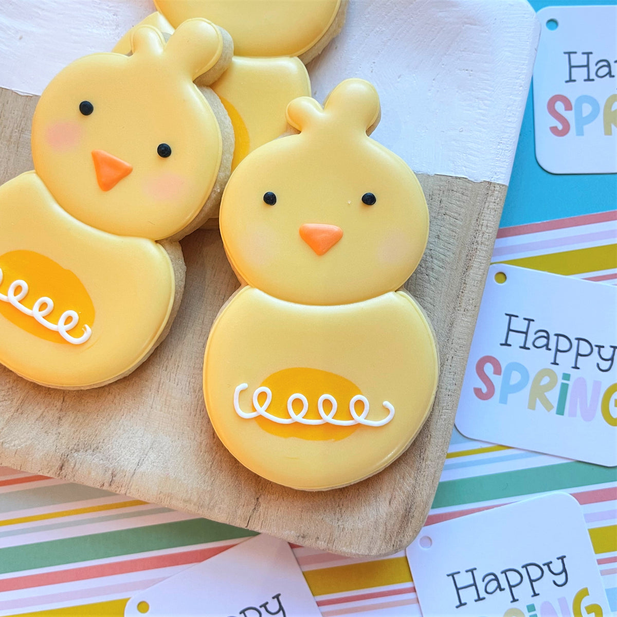 Yellow chick-shaped cookies with orange beaks and white icing swirls made using chick roly poly flour box club cookie cutter