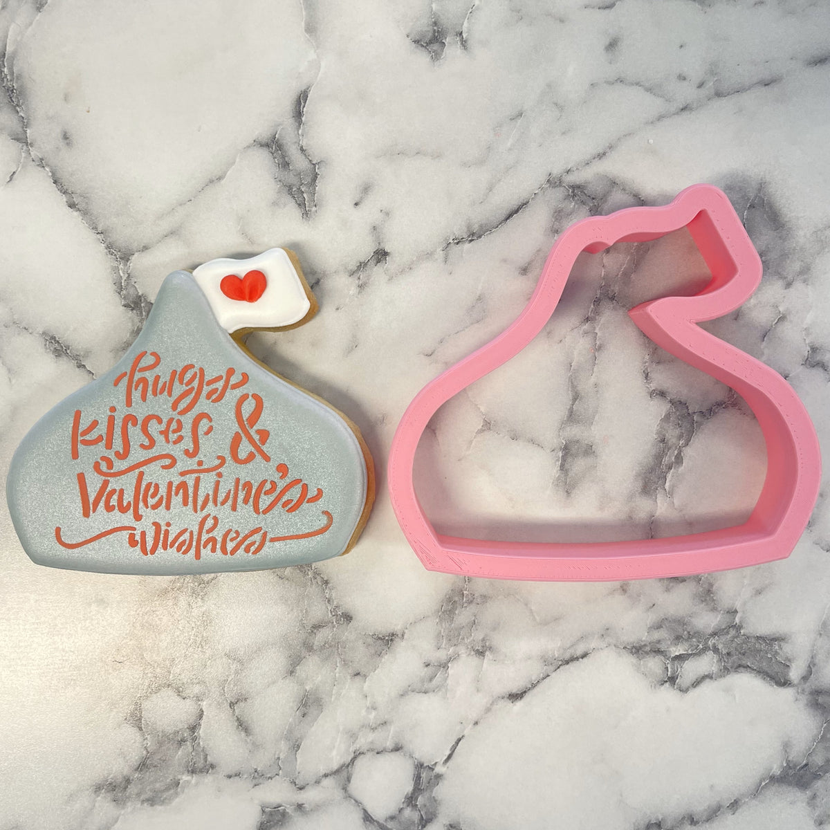 Pink silicone chocolate kiss cookie cutter next to a decorated gray sugar cookie with orange icing and red heart detail