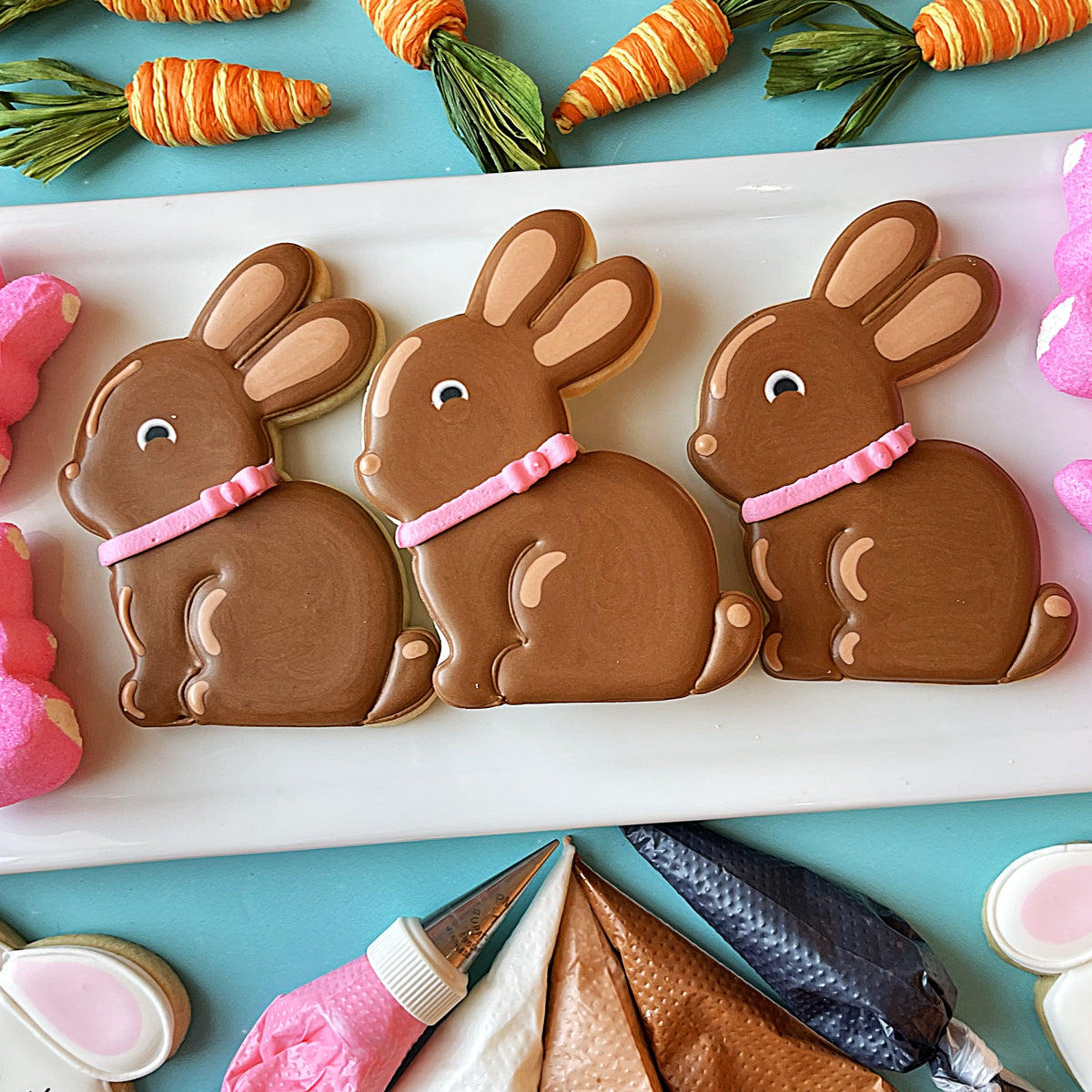 Three chocolate brown bunny-shaped cookies with pink icing collars and white eyes on a white plate for easter sweets online class recording