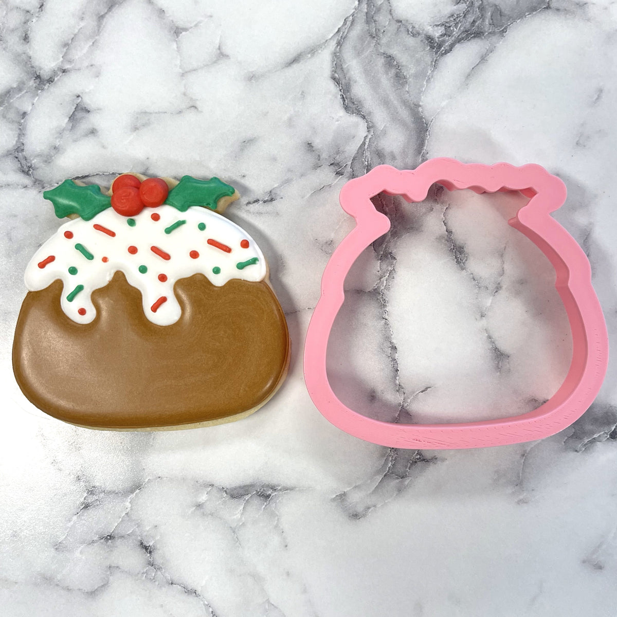 A pink plastic christmas pudding cookie cutter with a scalloped top edge beside a decorated pudding-shaped cookie