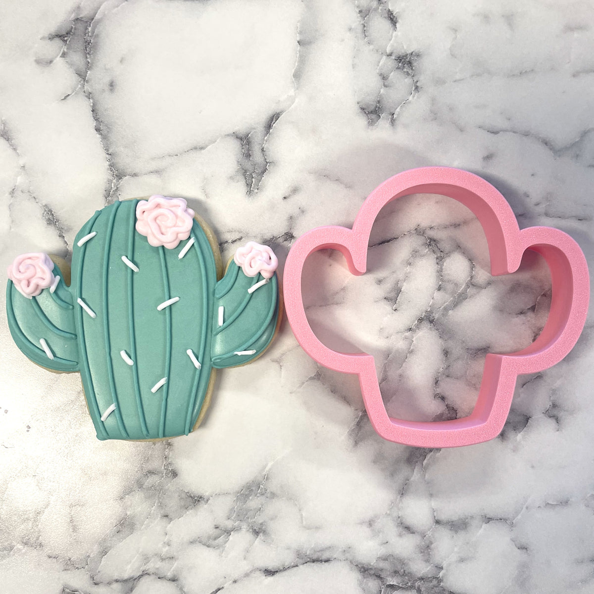 A pink plastic chubby cactus cookie cutter beside a teal frosted cactus sugar cookie with white spines and pink roses