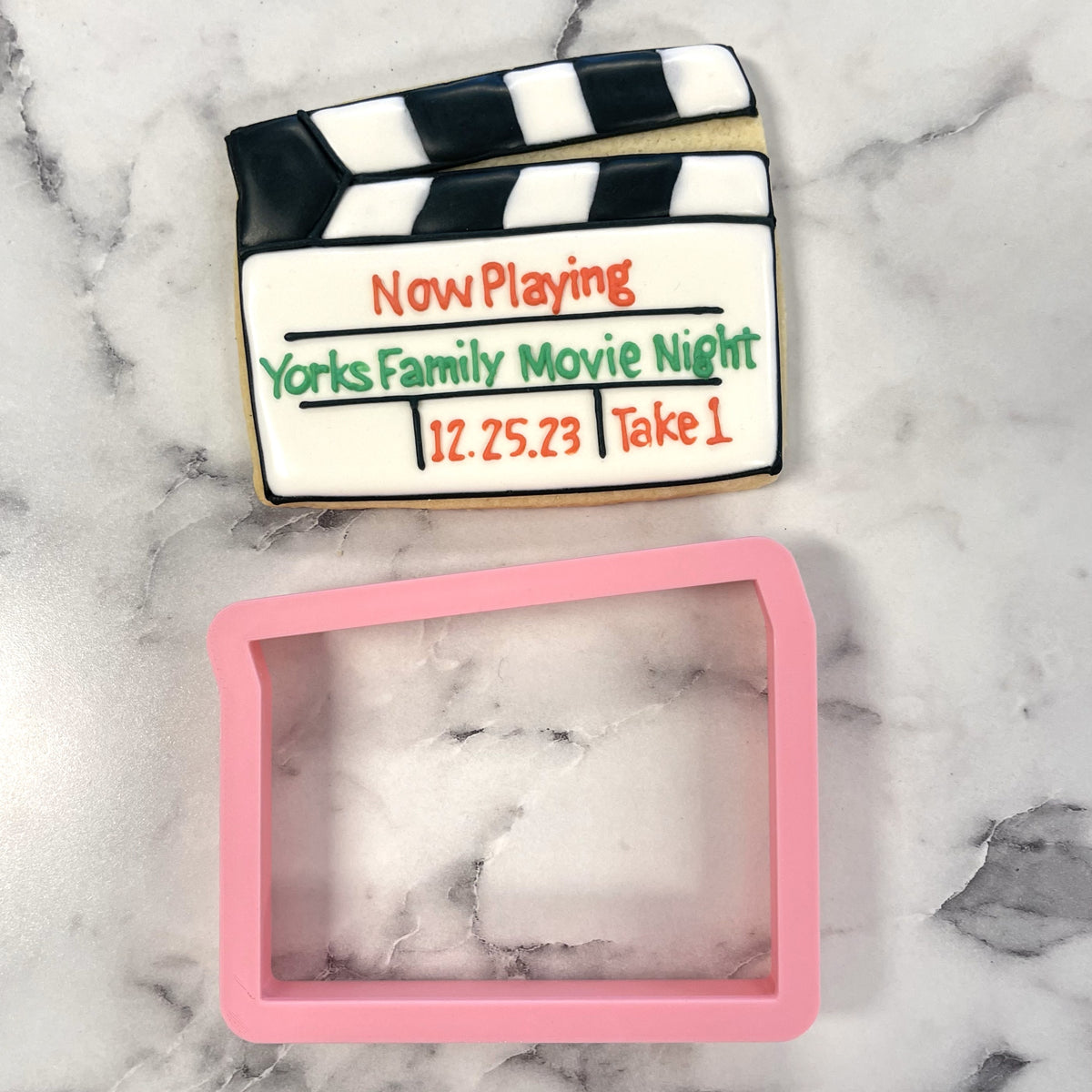 A decorated clapperboard cookie with black and white stripes featuring now playing in orange and yorks family movie night in green