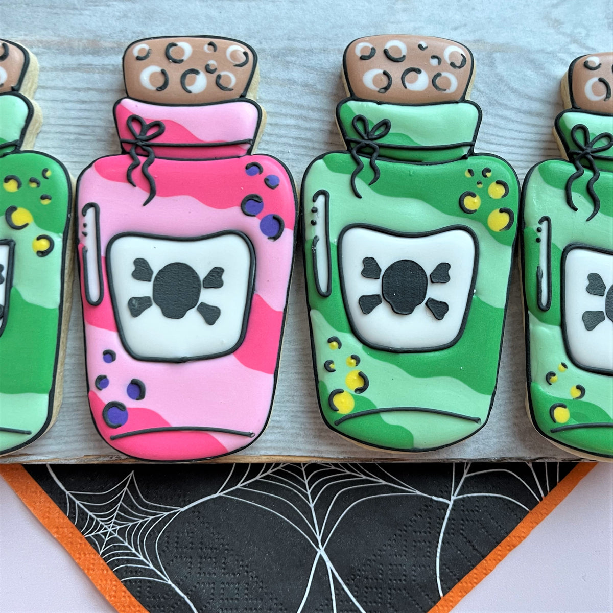 Pink and green camouflage sugar cookies shaped like potion bottles with black skull labels for comic halloween stencil