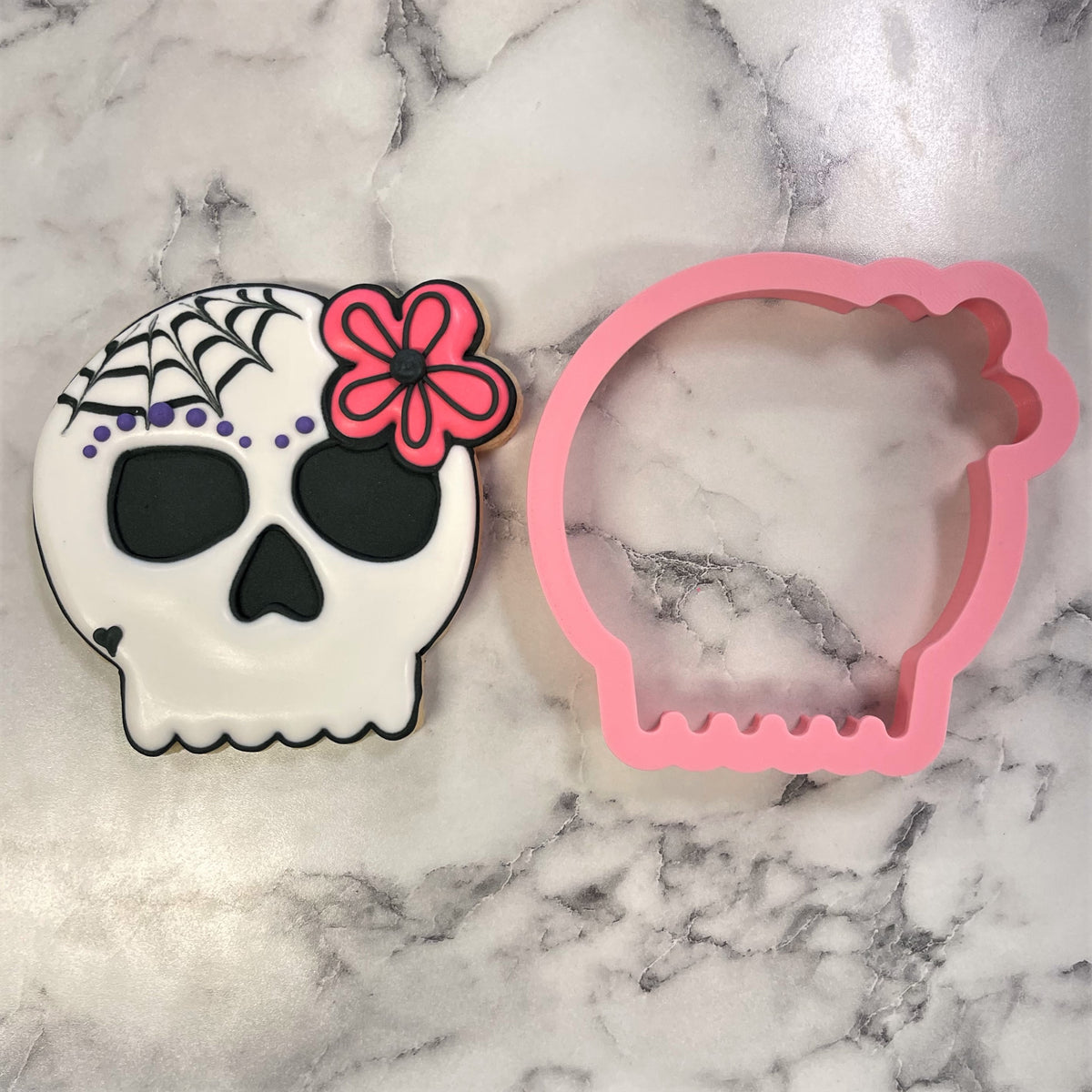 Comic skull girl cookie cutter pink silicone beside decorated white sugar skull cookie with black icing and purple dots
