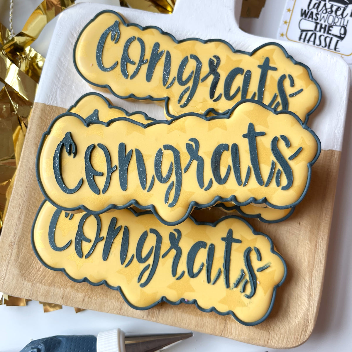 Yellow congrats cookies with dark blue glittery lettering and scalloped edges made using a congrats cookie cutter and stencil set