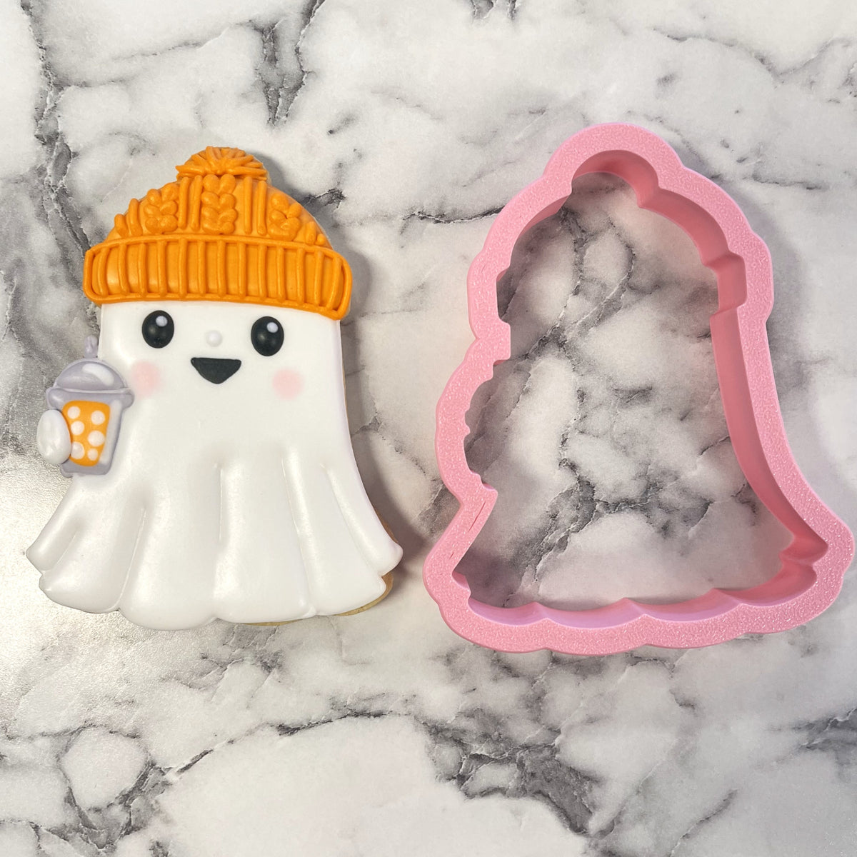 A white ghost-shaped cookie with an orange knitted beanie and drink beside a pink bell-shaped cozy ghost cookie cutter