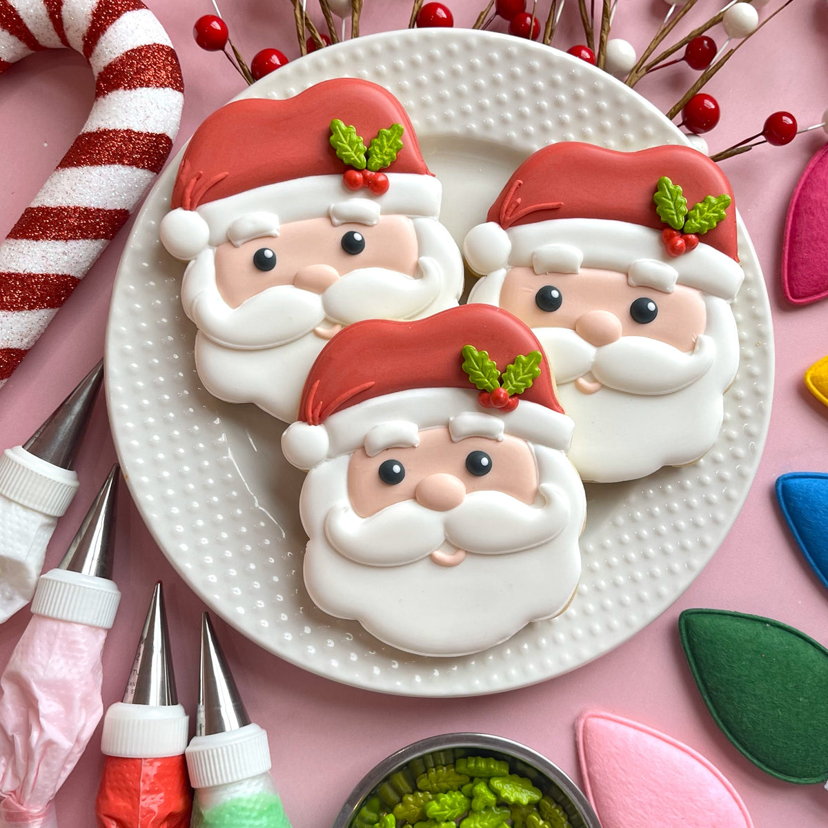 Three cute santa face cookies with red hats green holly white icing beards and detailed facial features made using a cookie cutter