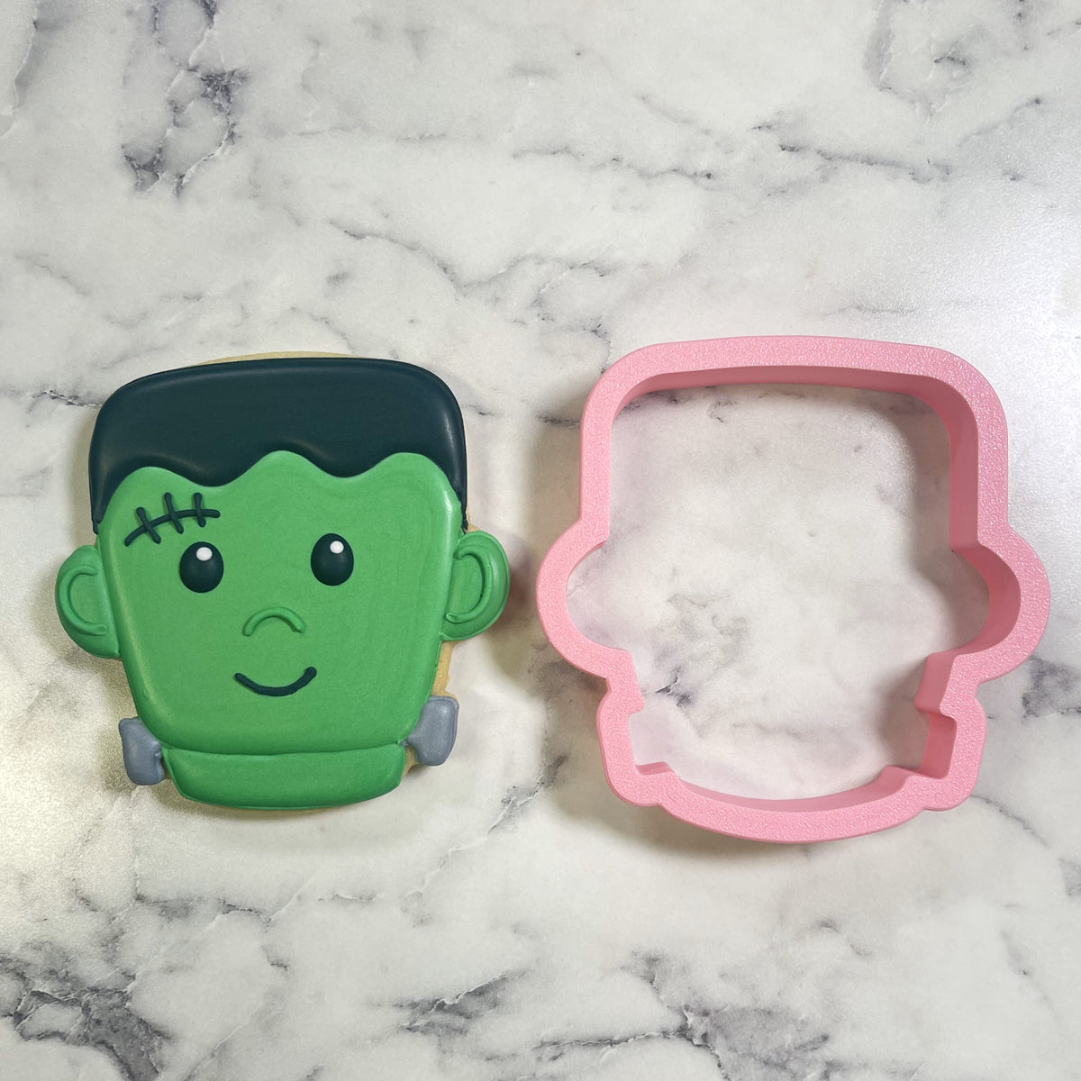 Cute chubby frank cookie cutter pink plastic next to a green icing decorated frankenstein-themed cookie