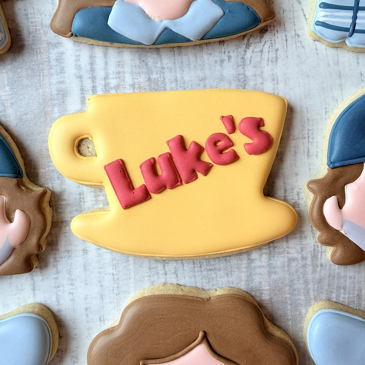A yellow frosted cookie shaped like a coffee cup with red luke’s icing made using a diner mug cookie cutter