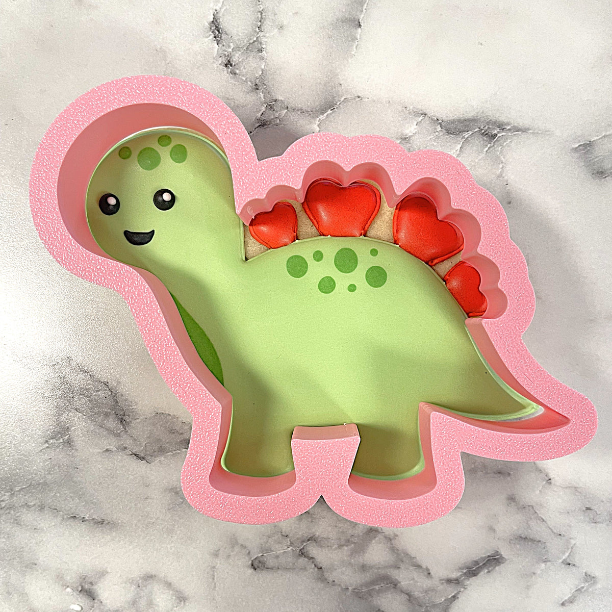 A pink foam dinosaur cookie cutter with green interior and red heart-shaped spikes for baking dinosaur cookies