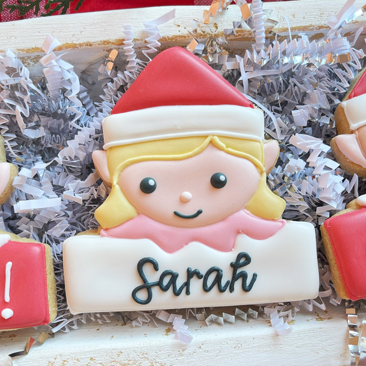A personalized sugar cookie shaped like a blonde elf in a red and white santa hat with the name sarah below