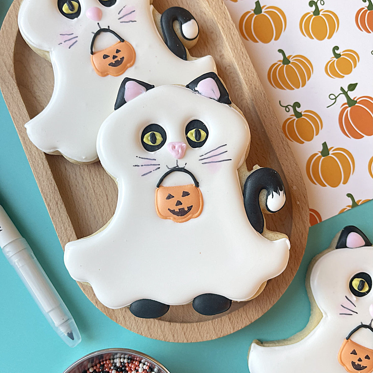 White frosted cat-shaped cookie with black ears green eyes pink nose and orange pumpkin candy bag for ghost pets cookie cutter bundle