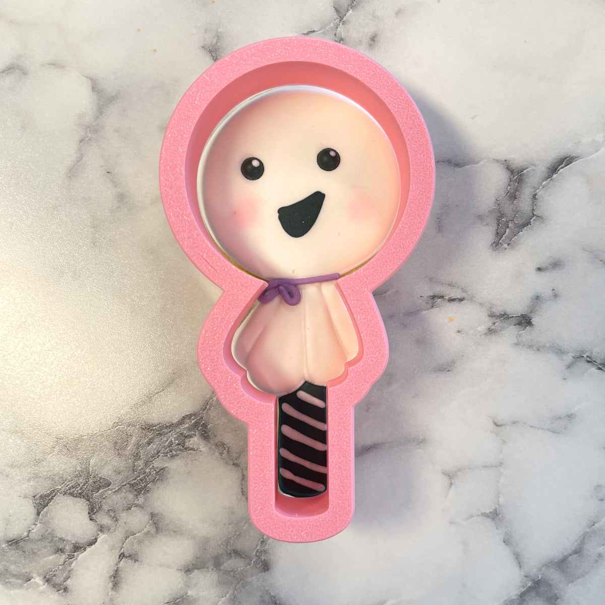 Pink silicone mold shaped like a smiling ghost lollipop with blush cheeks and a striped stick for cookie cutter use