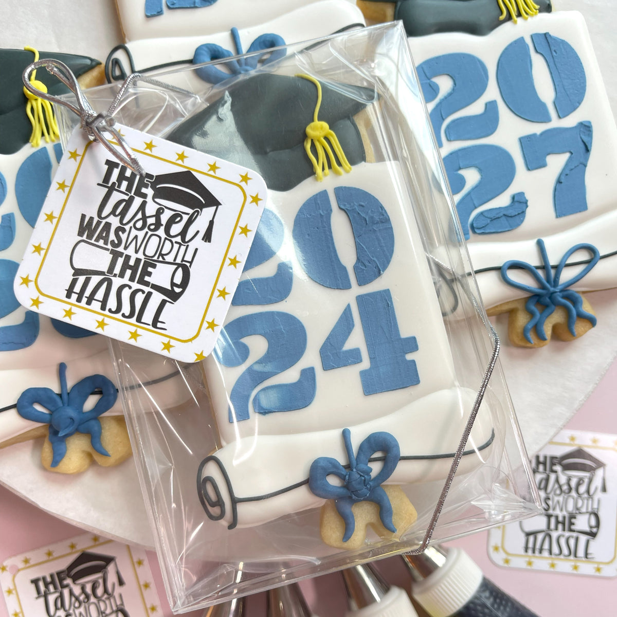 White graduation-themed cookie with blue 2024 icing and decorative blue ribbon made using grad cap yearly plaque cookie cutter and stencil