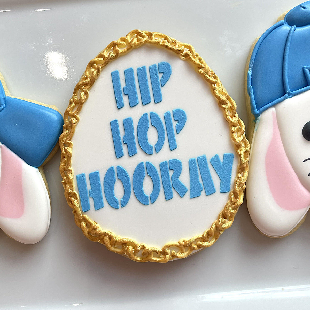 White sugar cookie shaped like an egg decorated with blue icing spelling hip hop hooray and a golden chain border made using 3.5 inch egg cookie cutter