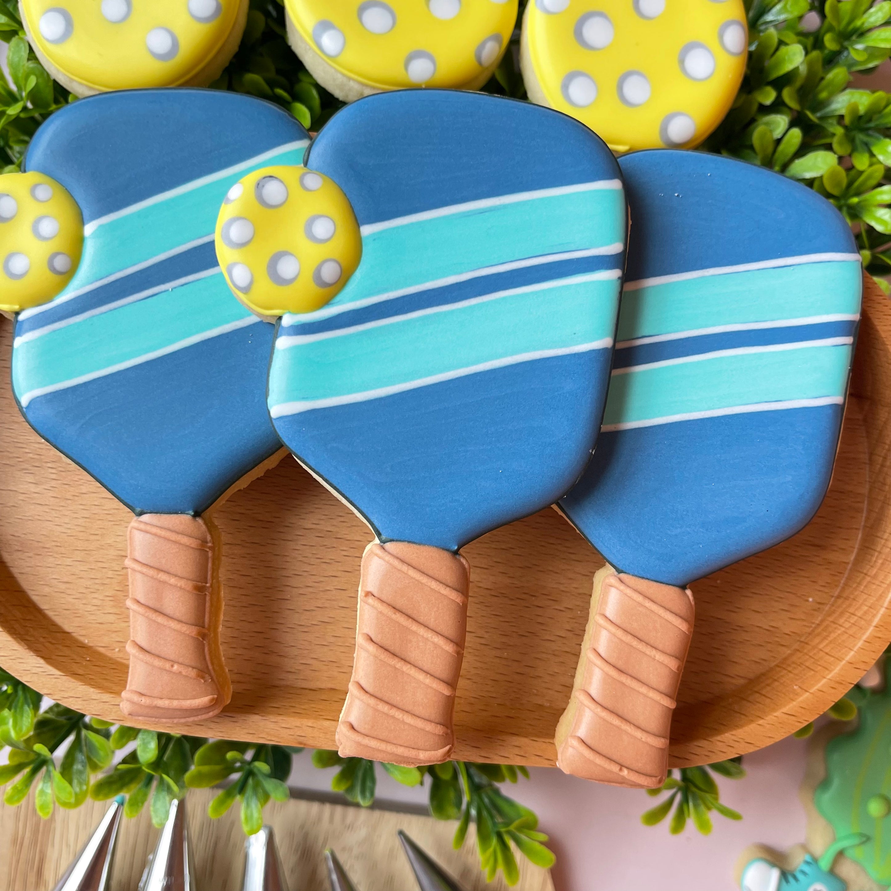 Blue and teal striped pickleball paddle-shaped cookies with yellow polka-dotted ball decorations made with a pickleball paddle with ball cookie cutter