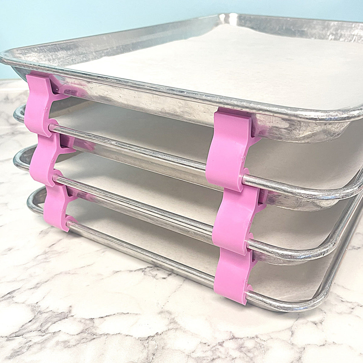 Stack of four silver metal baking trays held together by pink plastic clips from 24pc baking tray stacking spacer kit