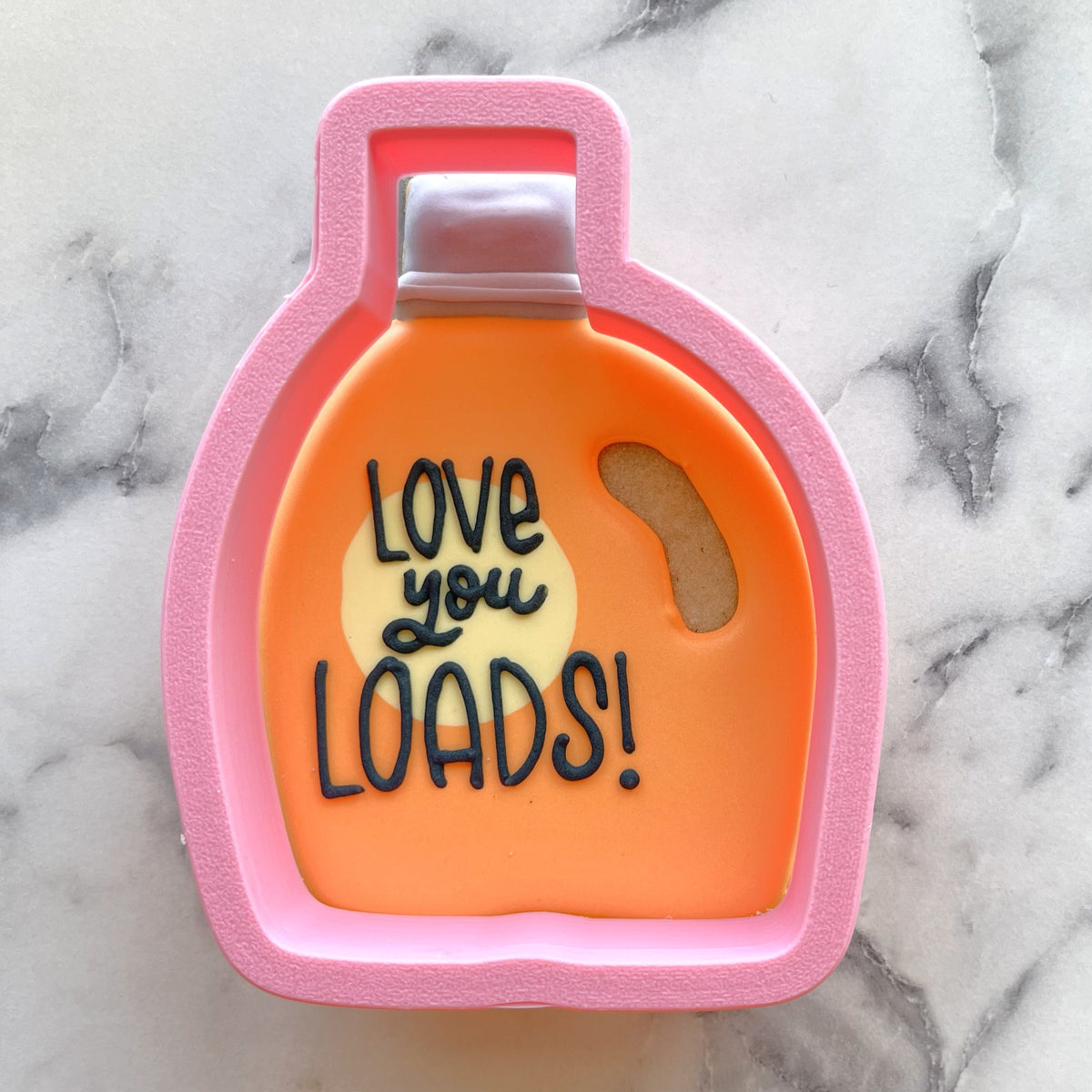 Pink silicone laundry detergent cookie cutter with orange interior and black icing reading love you loads