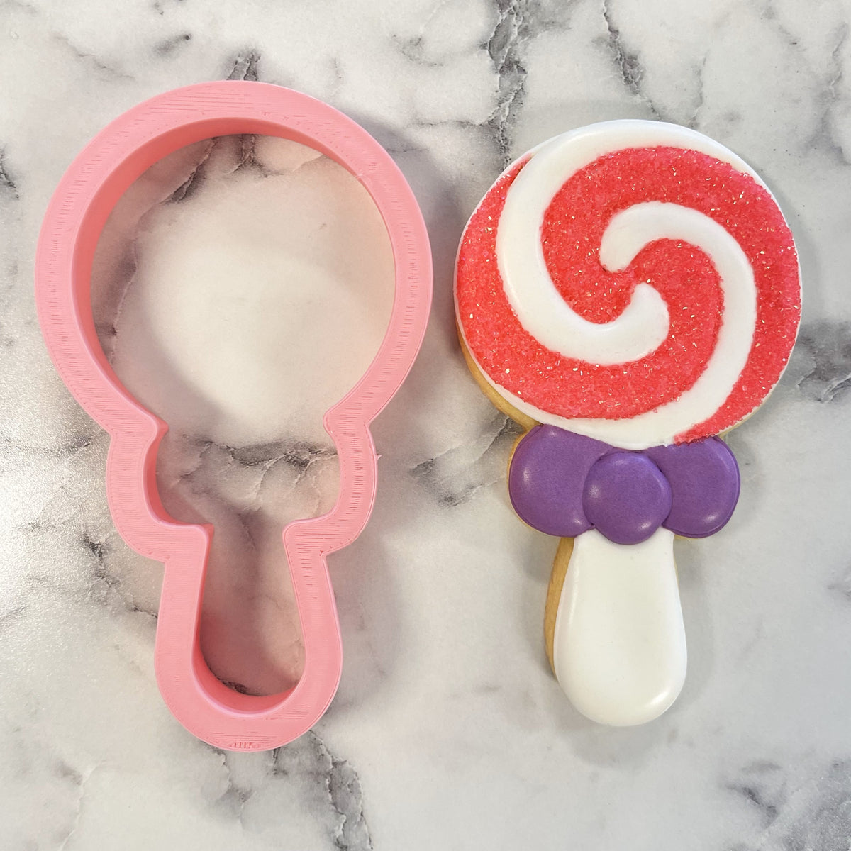 Pink plastic lollipop cookie cutter next to a red and white swirl sugar cookie with purple bow decoration