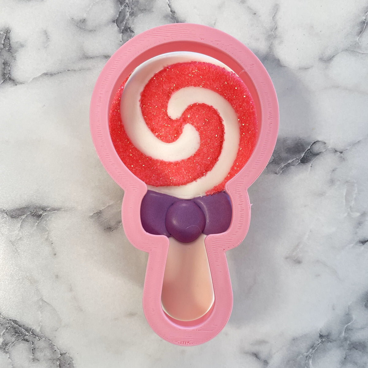 Pink lollipop cookie cutter bath bomb mold with a red and white swirled glitter interior and a purple bow detail