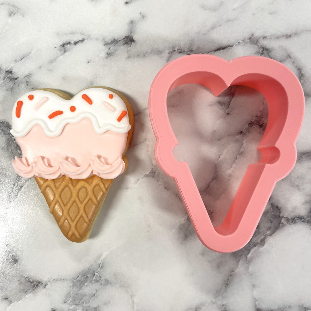 Mini heart ice cream cone cookie cutter pink silicone next to a heart-shaped sugar cookie with pink and white icing