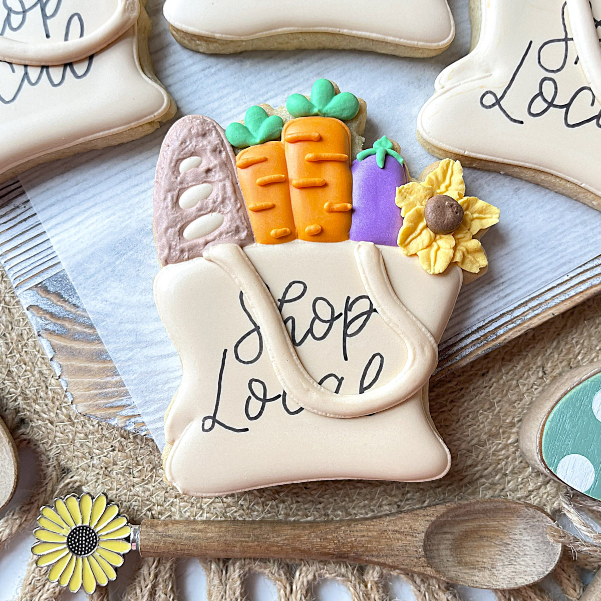 Cream-colored sugar cookie shaped like a shopping bag with shop local icing and vegetable decorations market tote flour box club cookie cutter