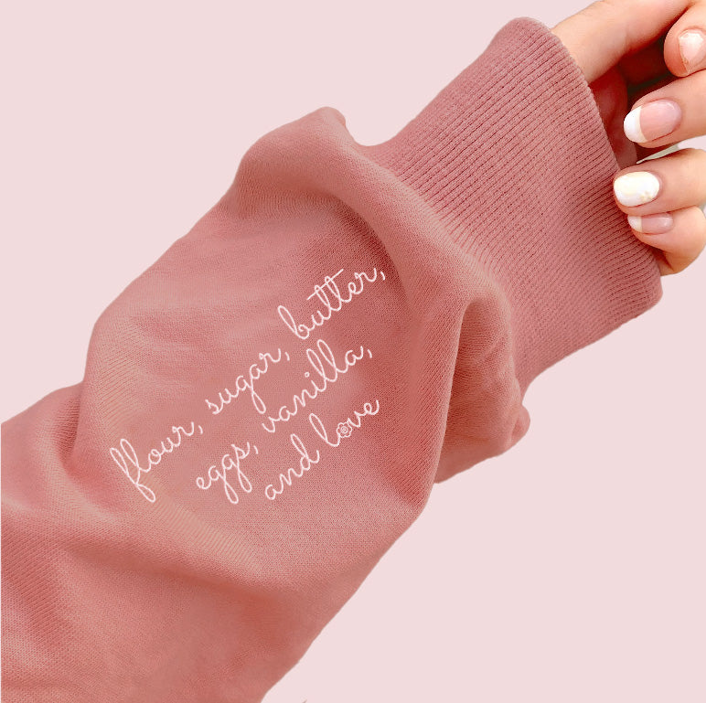 A soft pink long-sleeved sweatshirt with white cursive embroidery of baking ingredients on the sleeve in mauve