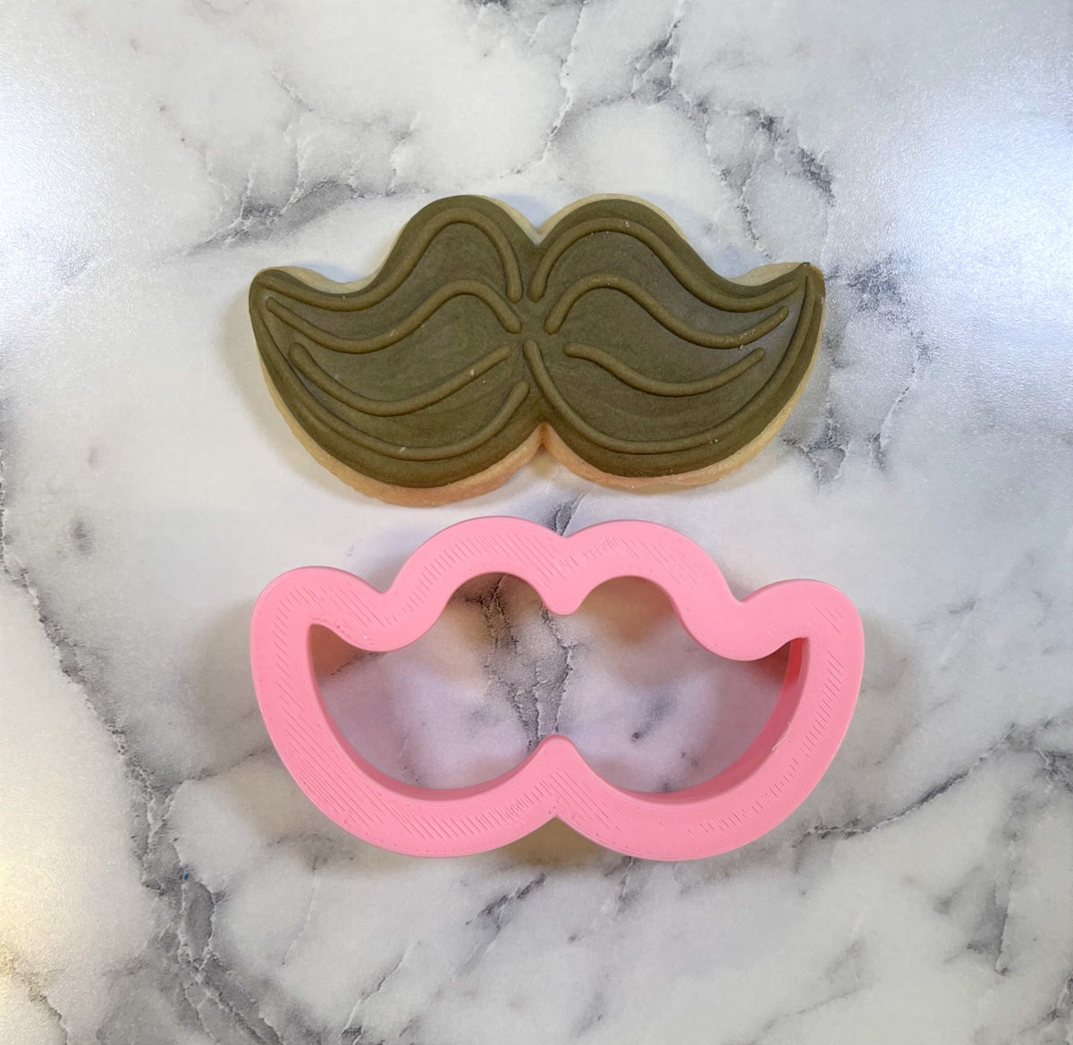 Mini mustache cookie cutter in pink plastic with detailed wavy design beside olive green mustache sugar cookie