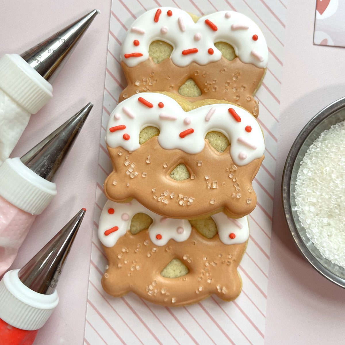 Mini pretzel cookie cutter shaped cookies with tan icing white frosting drizzle and red and pink sprinkles