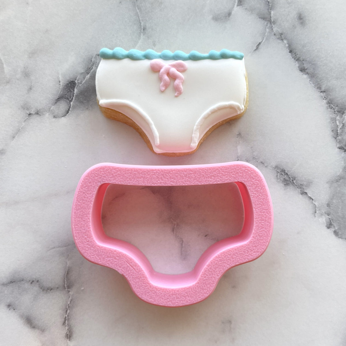 A pink silicone mini underpants cookie cutter shown with a matching decorated sugar cookie for baking use