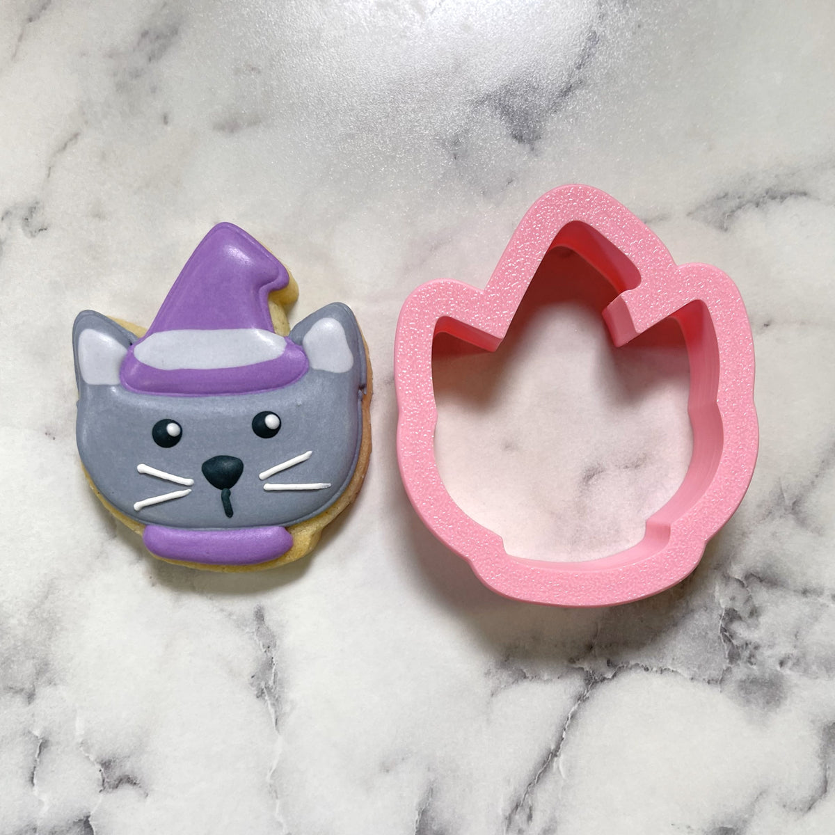 A pink cat-shaped mini cute chubby cat witch cookie cutter next to a gray witch-themed frosted sugar cookie