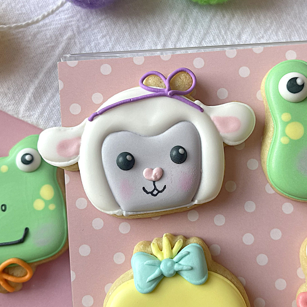 Mini spring lamb face cookie cutter with white frosted lamb cookie decorated with pink cheeks black eyes and purple bow