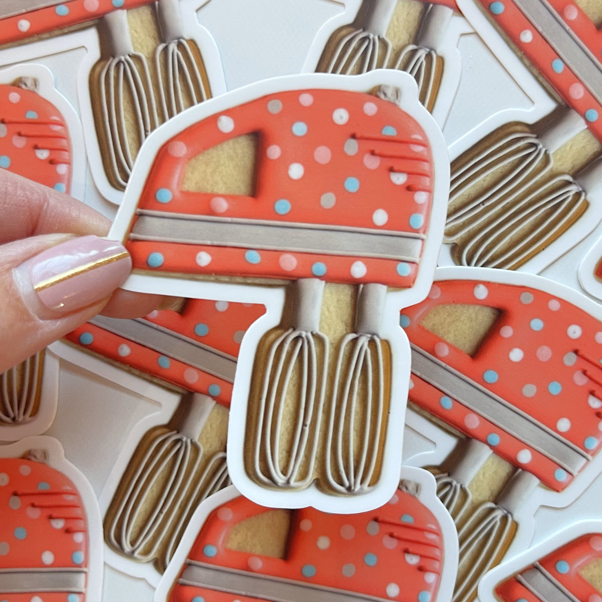 A hand holds a red polka-dotted stand mixer cookie sticker with beige whisks and silver accents for baking