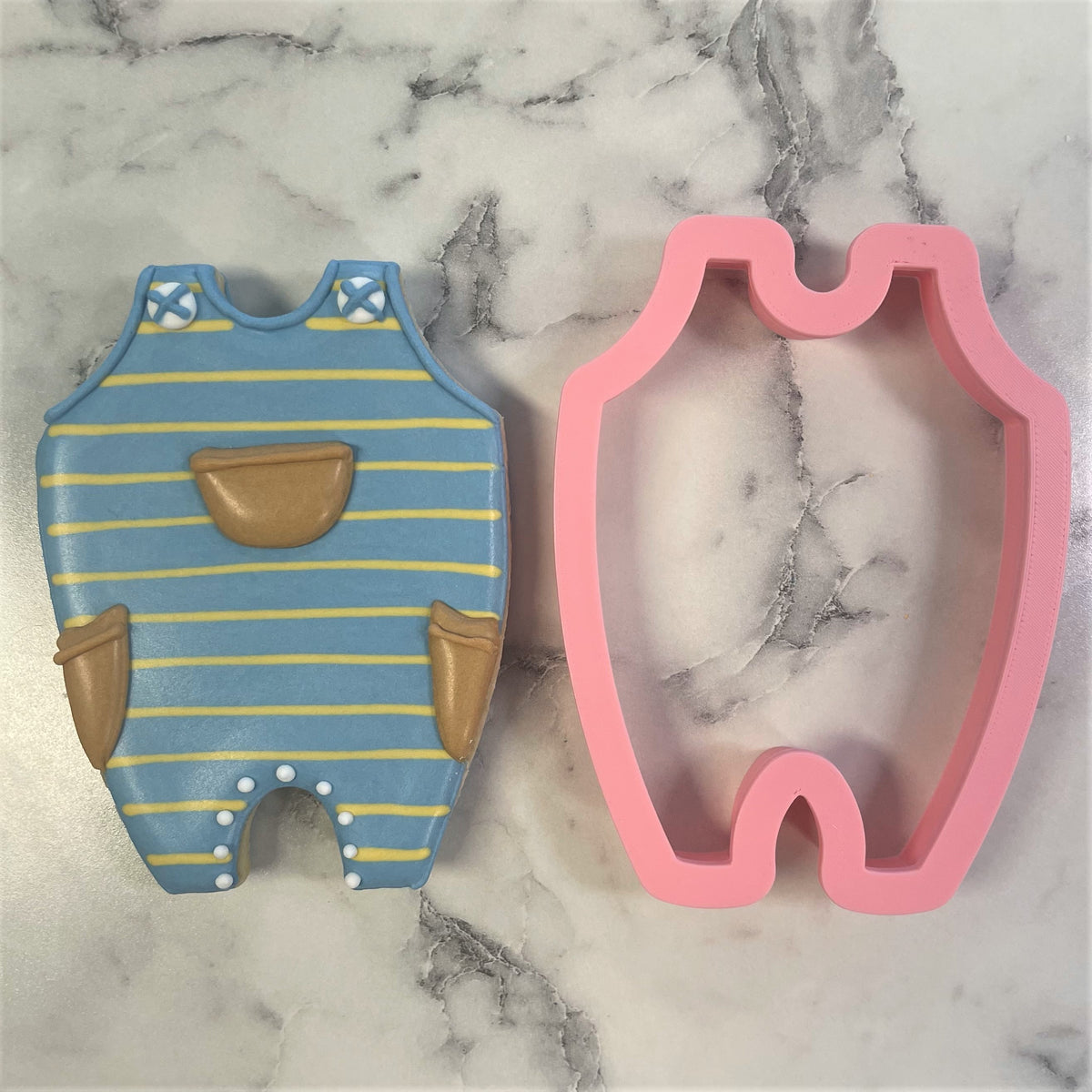 Blue and yellow striped baby overalls cookie with brown pockets and white polka dots beside its pink silicone mold