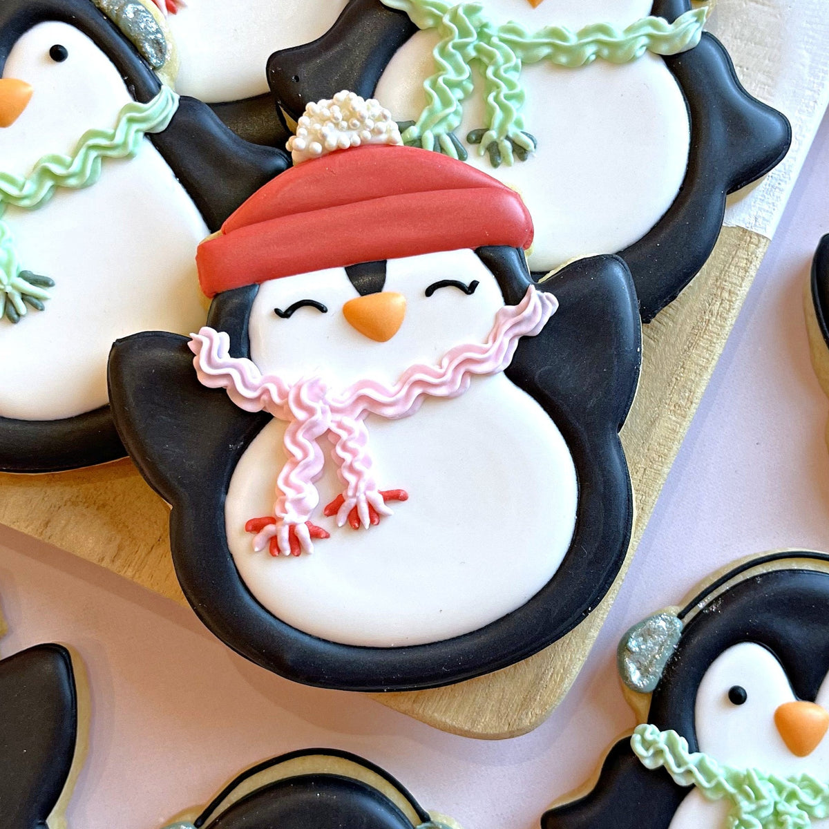 A penguin shaped sugar cookie decorated with black and white icing, red hat, pink scarf, and orange beak