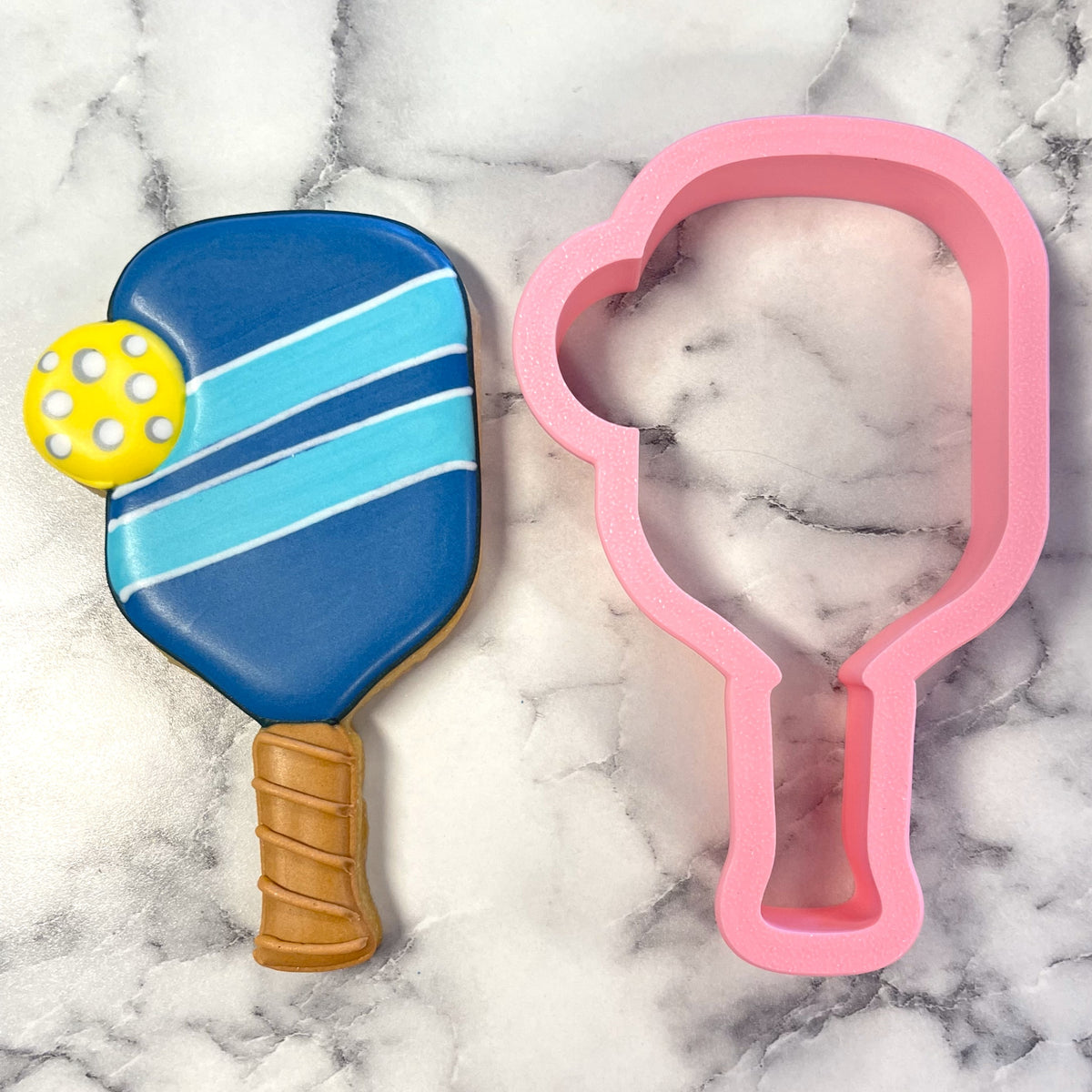 Blue and light blue striped pickleball paddle cookie with yellow ball detail next to pink silicone mold for cookie cutting