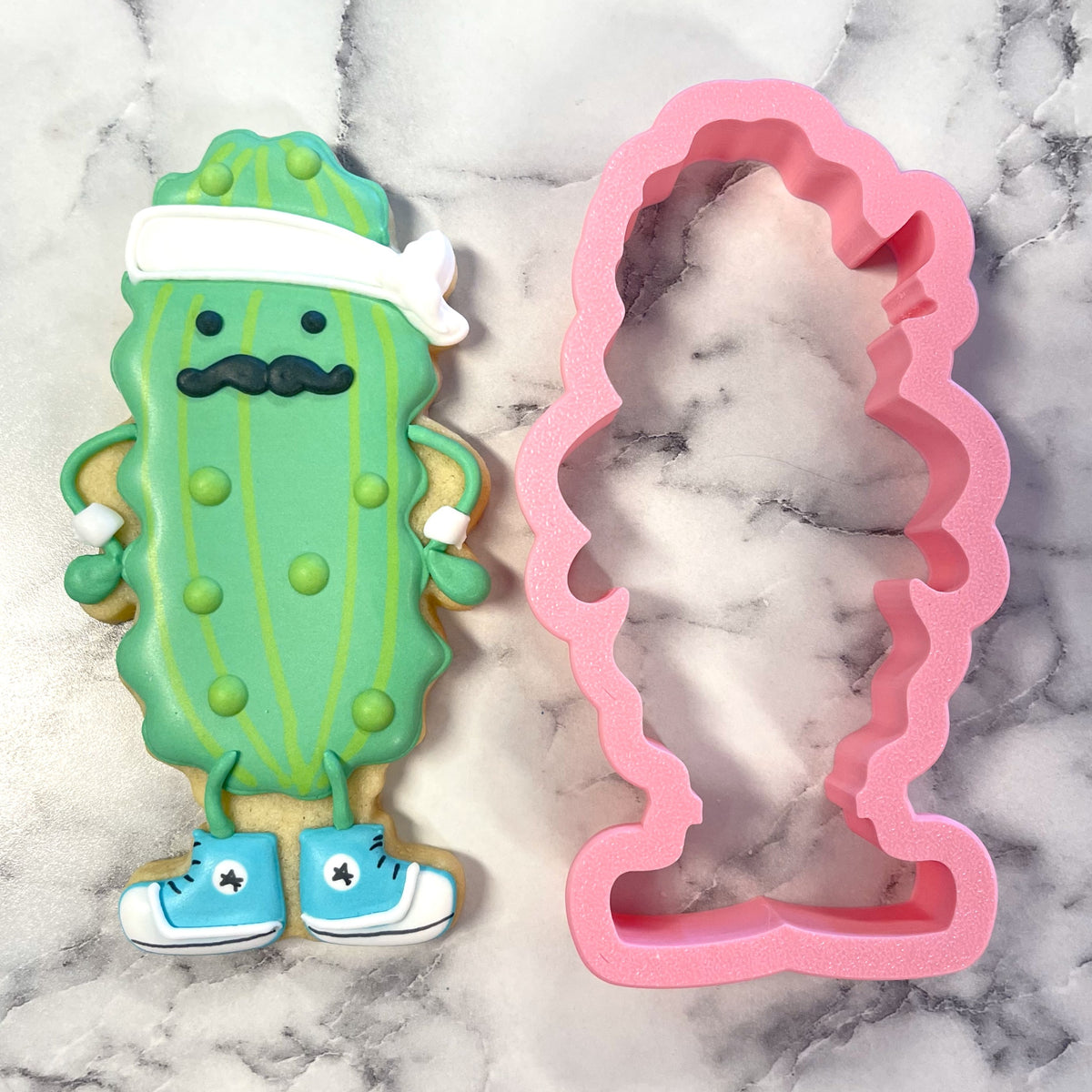 A whimsical green pickle baller cookie cutter shaped like a cookie with a white headband black mustache and blue sneakers