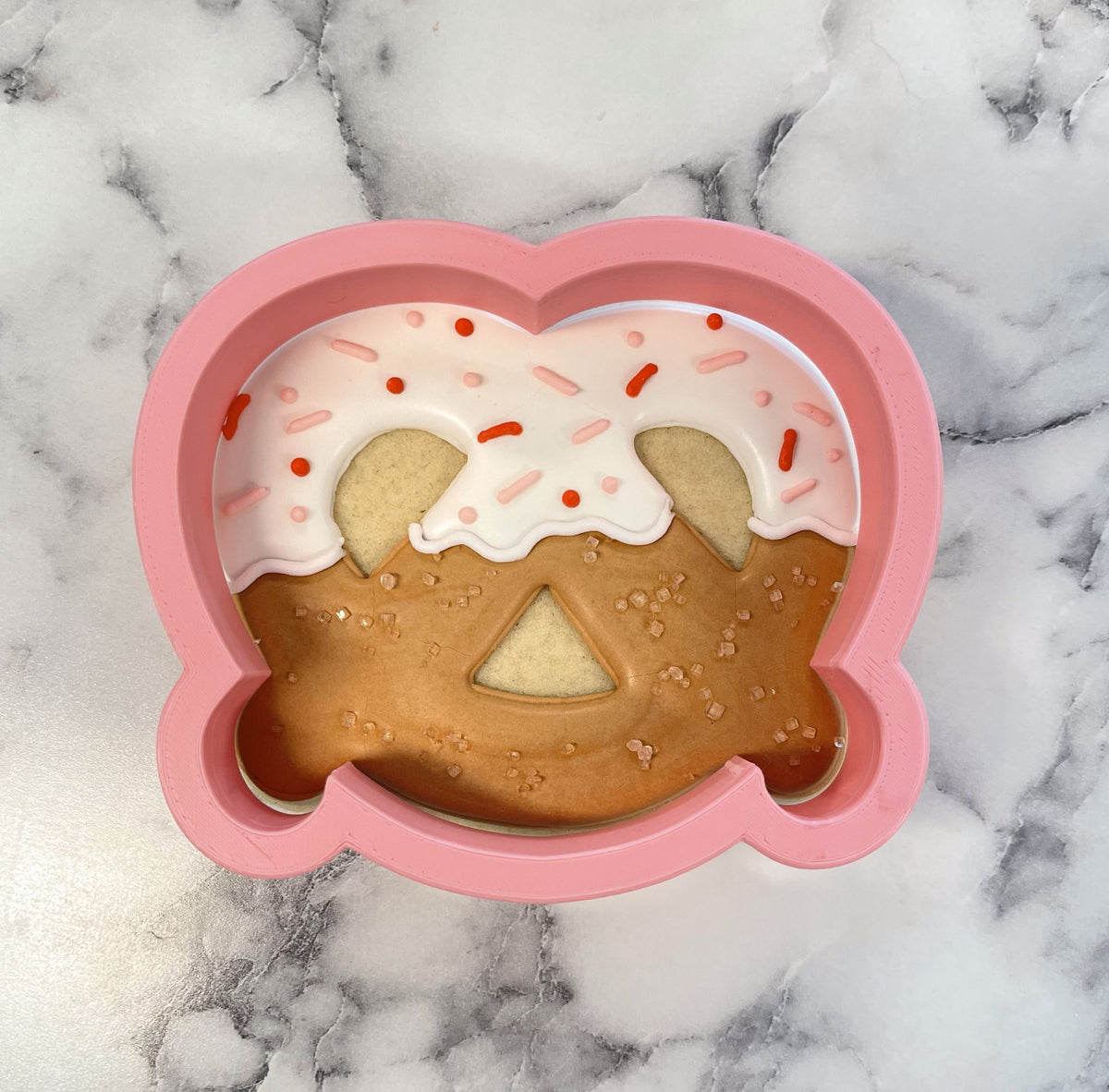 Pink silicone pretzel-shaped cookie cutter with white icing and red sprinkles over brown sugar coating for baking