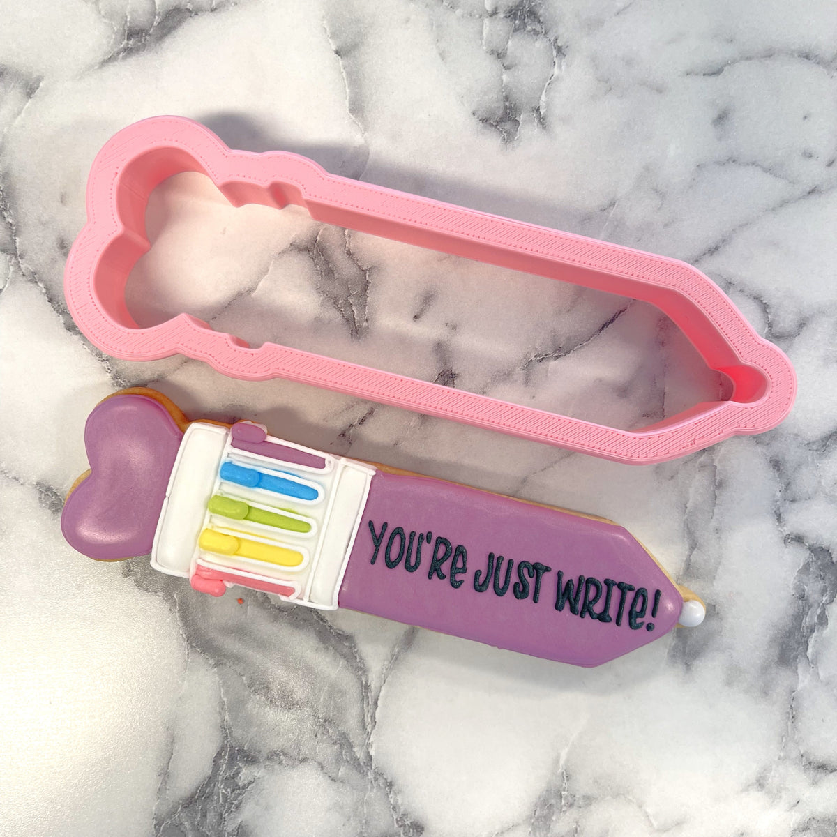 A pink bone-shaped retro click pen cookie cutter beside a purple pencil cookie with colorful keys and youre just write icing