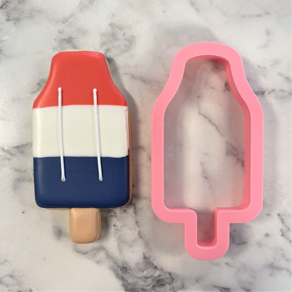 A pink silicone rocket pop flour box club cookie cutter next to a red white and blue striped popsicle cookie