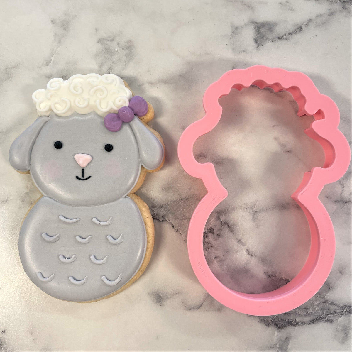 A pink silicone lamb roly poly flour box club cookie cutter with scalloped edge next to a decorated gray sugar cookie