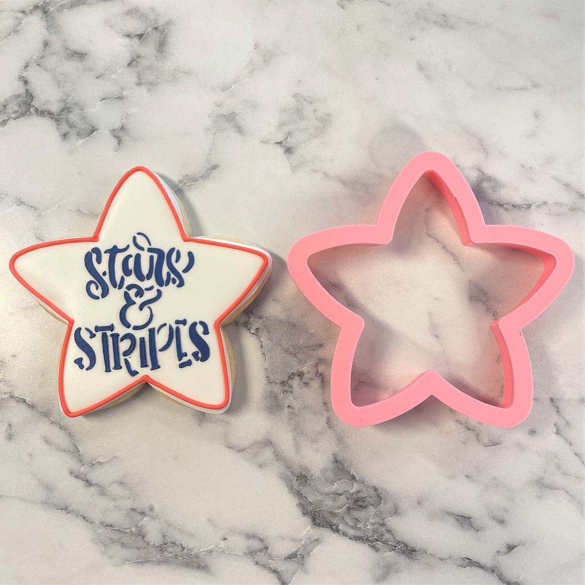 A pink star-shaped rounded star flour box club cookie cutter beside a white sugar cookie with blue icing and red border stars & stripes design