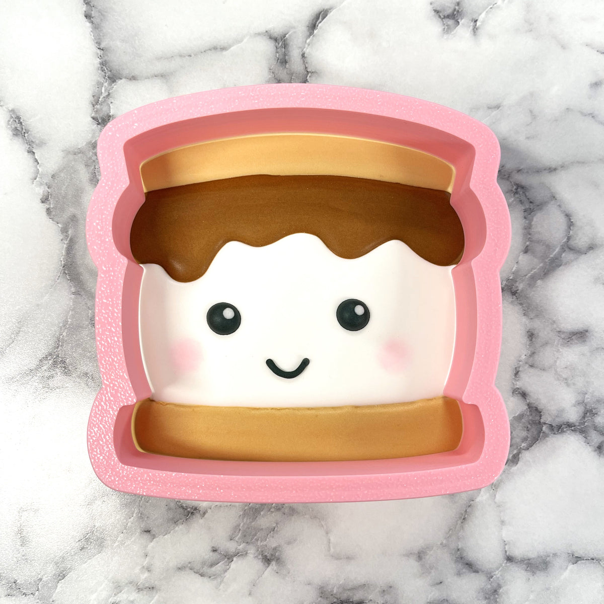 Pink silicone s’more cookie cutter shaped like a smiling s’more with white marshmallow chocolate and graham cracker edges