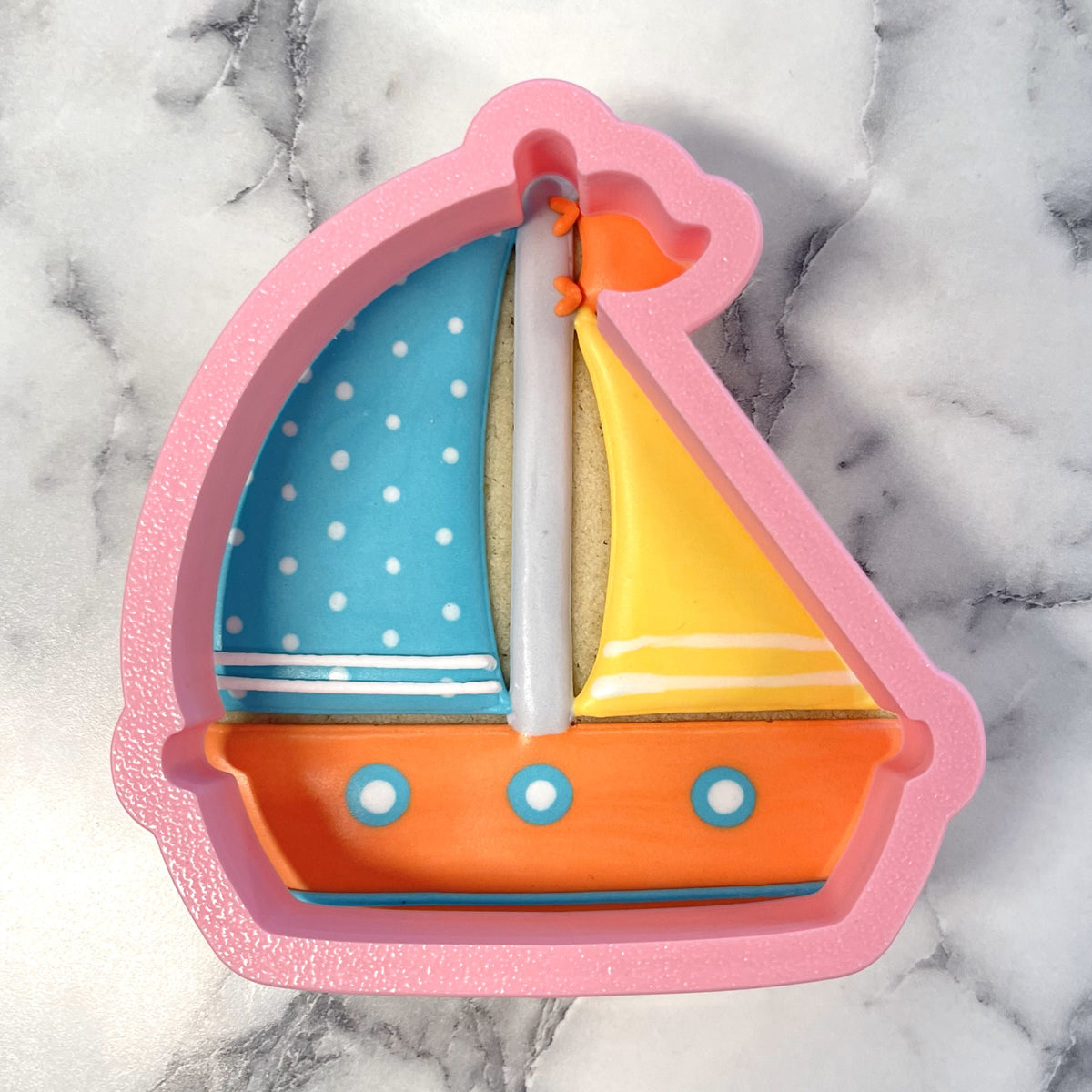 A pink silicone sailboat cookie cutter with blue and yellow sails orange hull and white polka dots